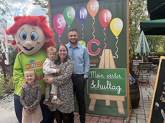 JOSKA Maskottchen Joschi begrüßt eine Familie mit zwei Kindern vor der bunten Schulanfangs-Fotowand im JOSKA Glasparadies.