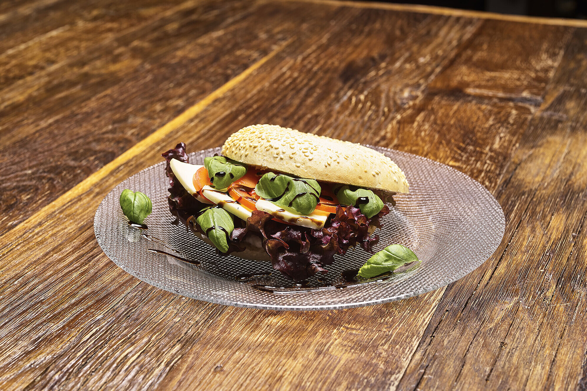 Vegetarisches Sandwich mit Käse, Tomate, Blattsalat und Basilikumblättern, garniert mit Balsamicocreme auf einem Glasteller.