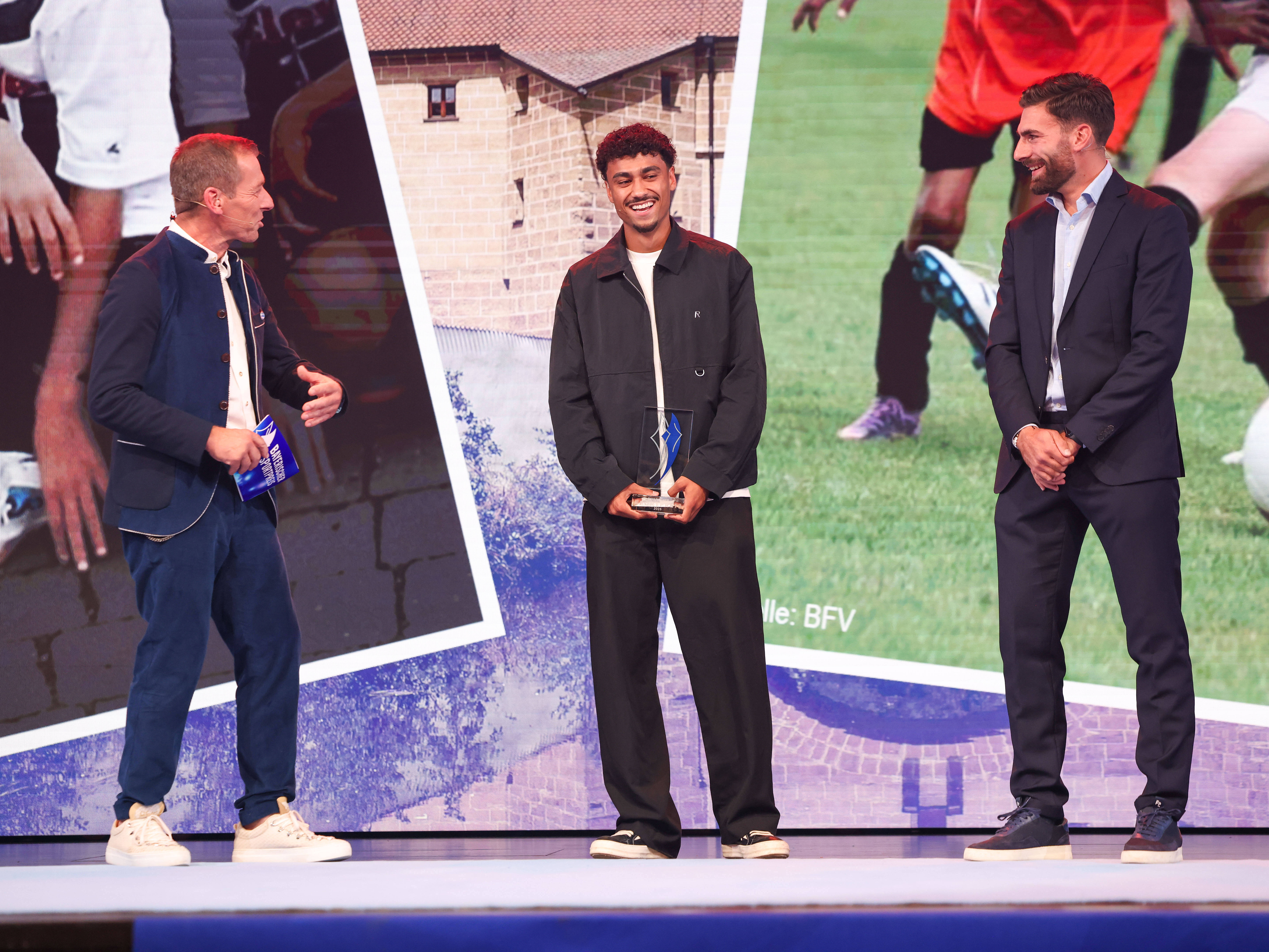 Ausgezeichneter Sportler mit gläserner Trophäe beim Bayerischen Sportpreis 2026 im Gespräch mit zwei Moderatoren auf der Bühne.