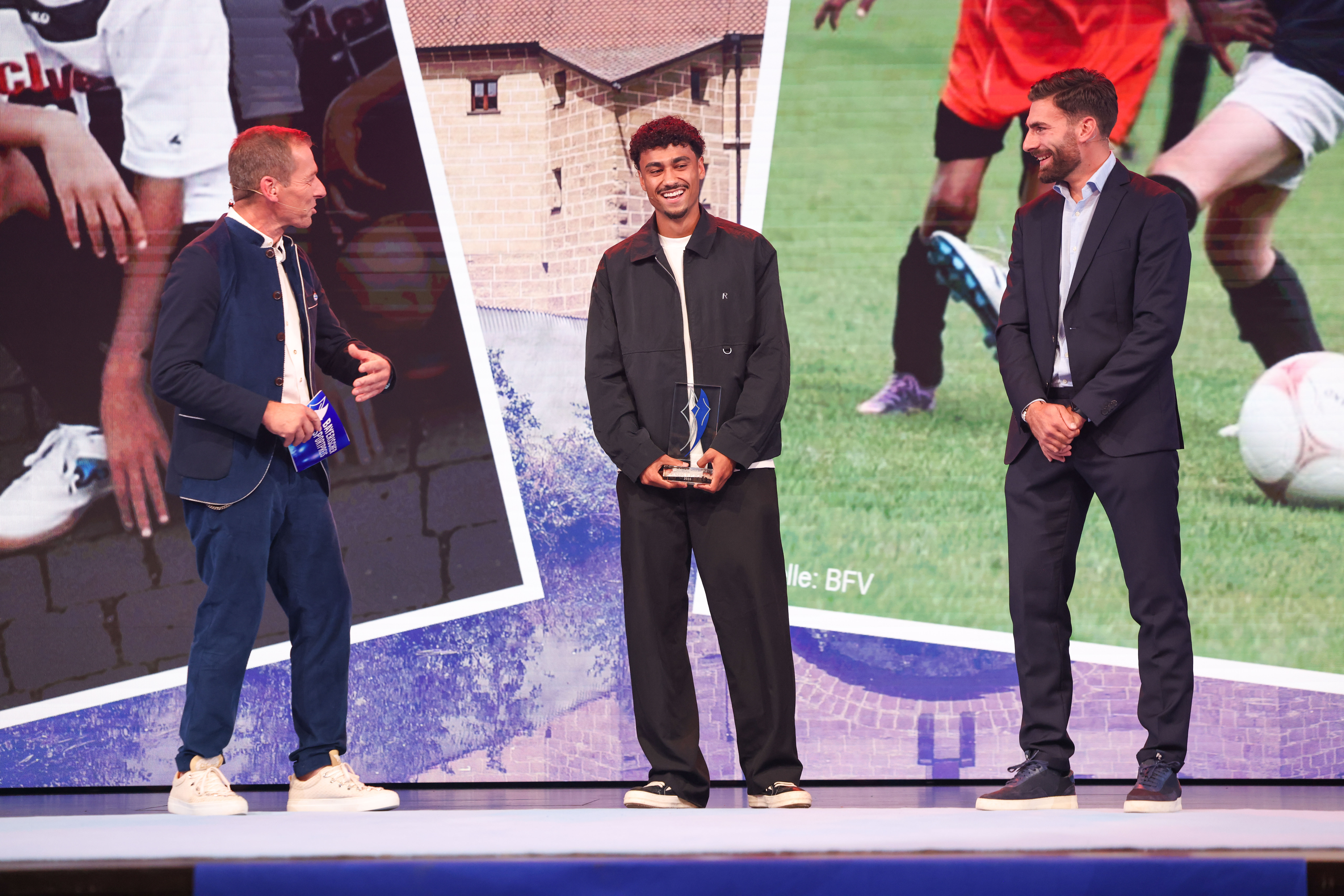 Ausgezeichneter Sportler mit gläserner Trophäe beim Bayerischen Sportpreis 2026 im Gespräch mit zwei Moderatoren auf der Bühne.